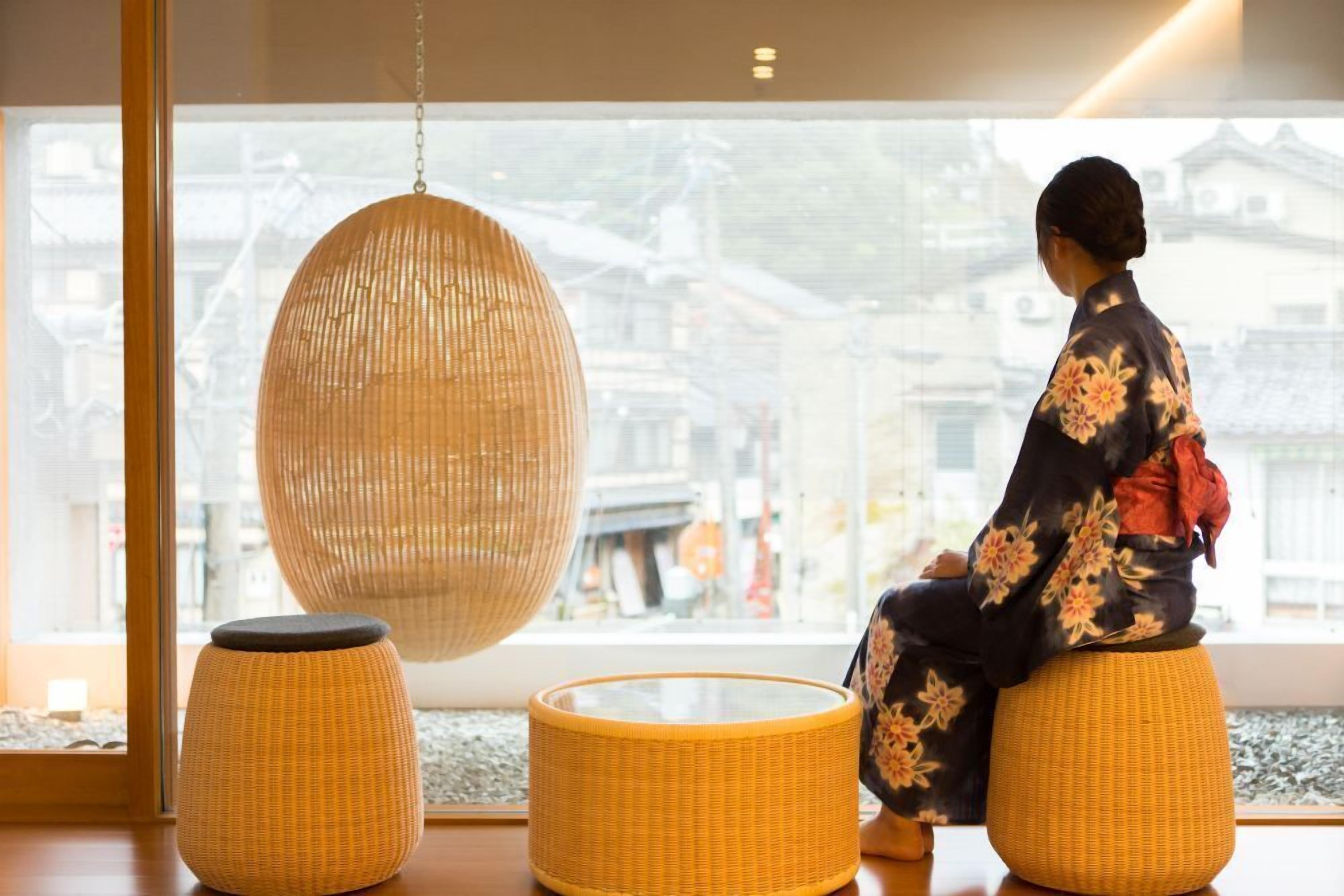 A room with a large window, a hanging wicker lamp, and a person sitting on a wicker stool.