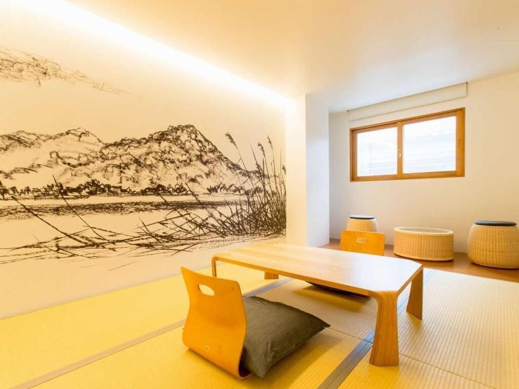 A room with tatami flooring, a low wooden table, a cushion, a wall-mounted artwork, and a window with blinds.