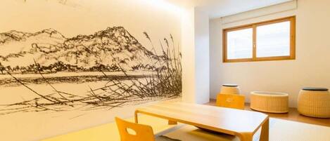 A room with tatami flooring, a low wooden table, a cushion, a wall-mounted artwork, and a window with blinds.