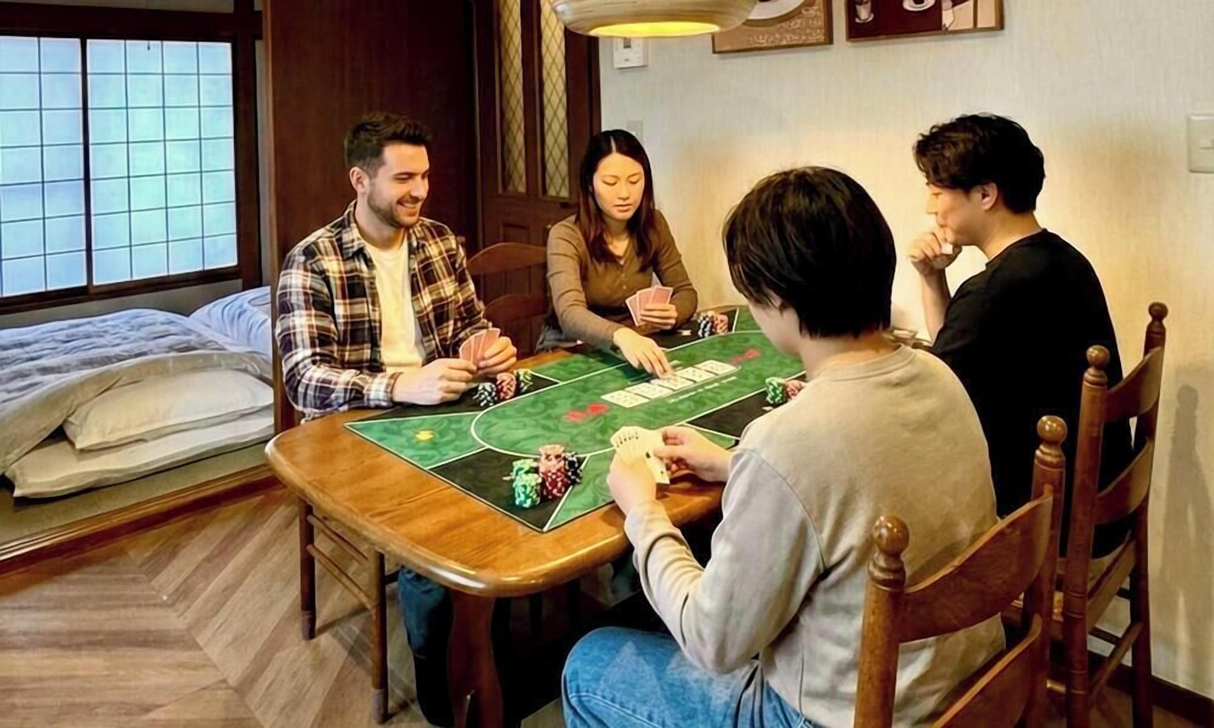 A group of people playing cards at a wooden table with a green felt tablecloth, in a room with a bed and a window.