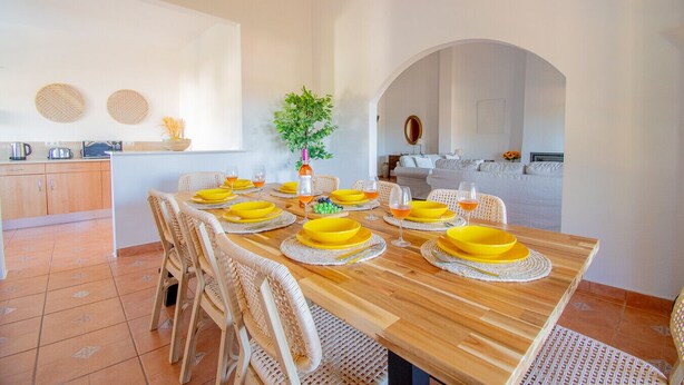 Un comedor con una mesa de madera dispuesta para seis personas, rodeada de sillas de mimbre, una cocina al fondo y un amplio arco.