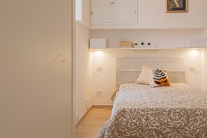 Un dormitorio con cama, cabecera, repisa con objetos y una lámpara fijada a la pared.