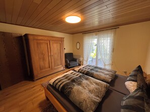 Une pièce avec un plafond en bois, une grande armoire, un lit avec une literie à motifs, une chaise et une fenêtre ornée de rideaux légers.