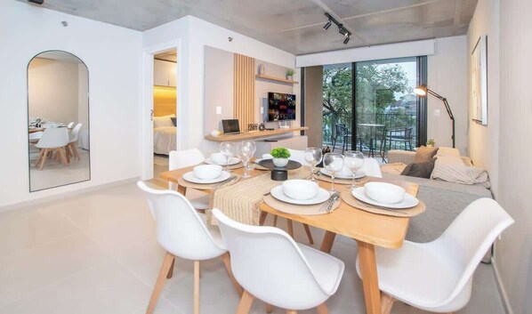 A modern dining area with a wooden table set for four, white chairs, and a sofa in the background. - Corporate Stays First del Sol (Asunción)