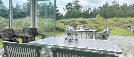 A covered outdoor deck with a table and chairs, overlooking a grassy area with trees.