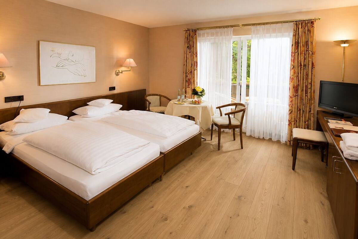 Ein Hotelzimmer mit zwei Betten, einem Schreibtisch mit Fernseher, einem Sessel und einem Tisch mit Blumenvase.
