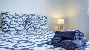 A bed with a patterned bedspread, a stack of folded towels, and a bedside lamp. - The Van Gogh House: 3 bedrooms, 4 beds, and easy parking (Tournefeuille)