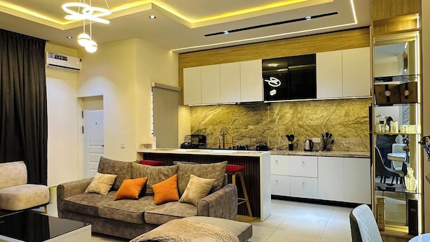 A modern living room with a sofa, ottoman, and a dining area with a table and chairs. - Chennad Apartment  (Abuja)