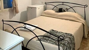 A single bed with a metal frame, a bedside table, a small cabinet, and a window with curtains. - Camere matrimoniali comunicanti (Sardegna)