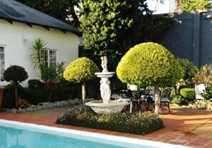 A pool with a fountain, surrounded by neatly trimmed hedges and a patio area with outdoor furniture. - Brooks Cottage (Pretoria)