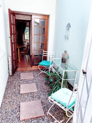 A narrow outdoor corridor with a wooden door, a metal chair with a blue cushion, and a small table with a plant. - Brooks Cottage (Pretoria)