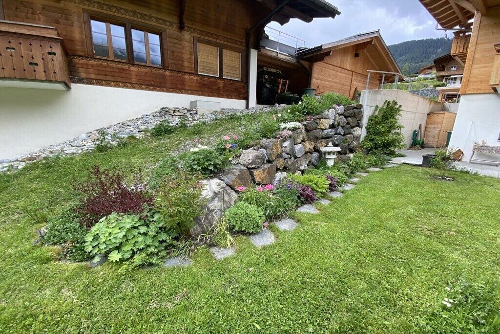Un chalet en bois avec un mur de soutènement en pierre, une cour gazonnée et une vue sur les montagnes en arrière-plan.