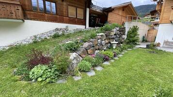 Un chalet en bois avec un mur de soutènement en pierre, une cour gazonnée et une vue sur les montagnes en arrière-plan.