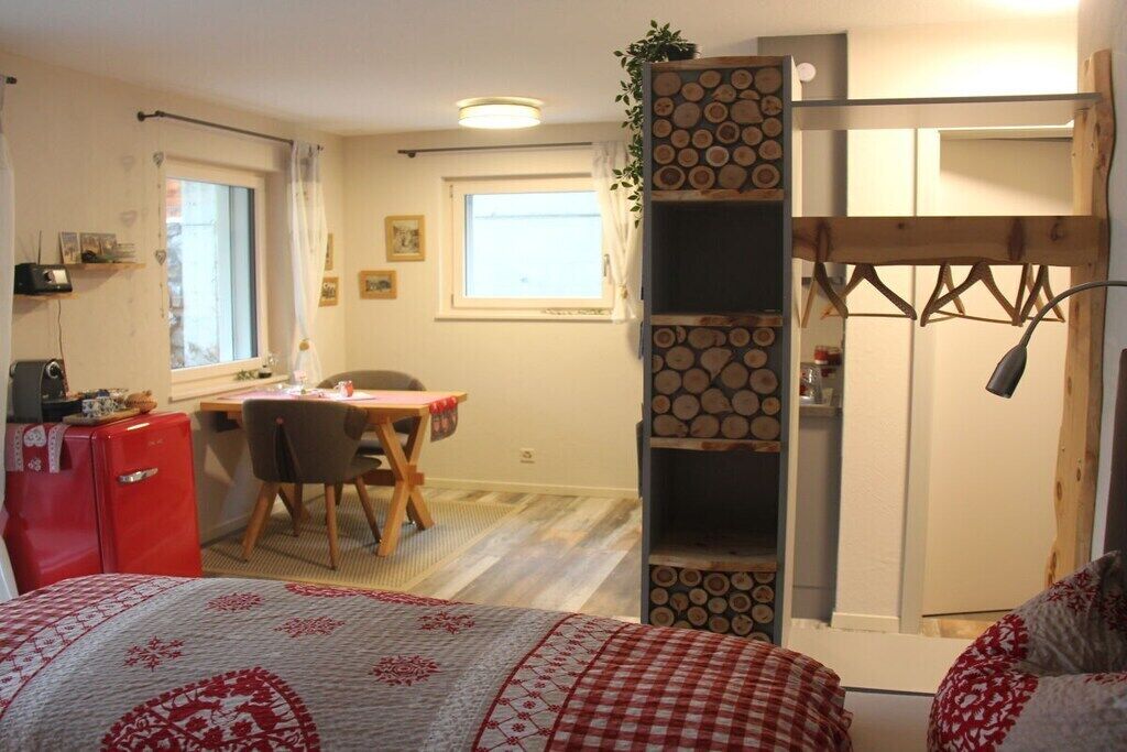Une petite pièce comprenant un lit, un bureau, une chaise, un réfrigérateur rouge et une étagère en bois avec des bûches.