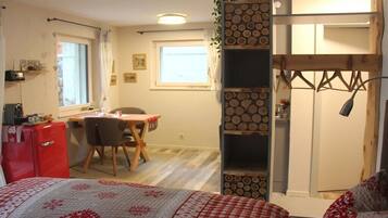 Ein kleines Zimmer mit einem Bett, einem Schreibtisch, einem Stuhl, einem roten Kühlschrank und einem Holzregal mit Holzscheiten.