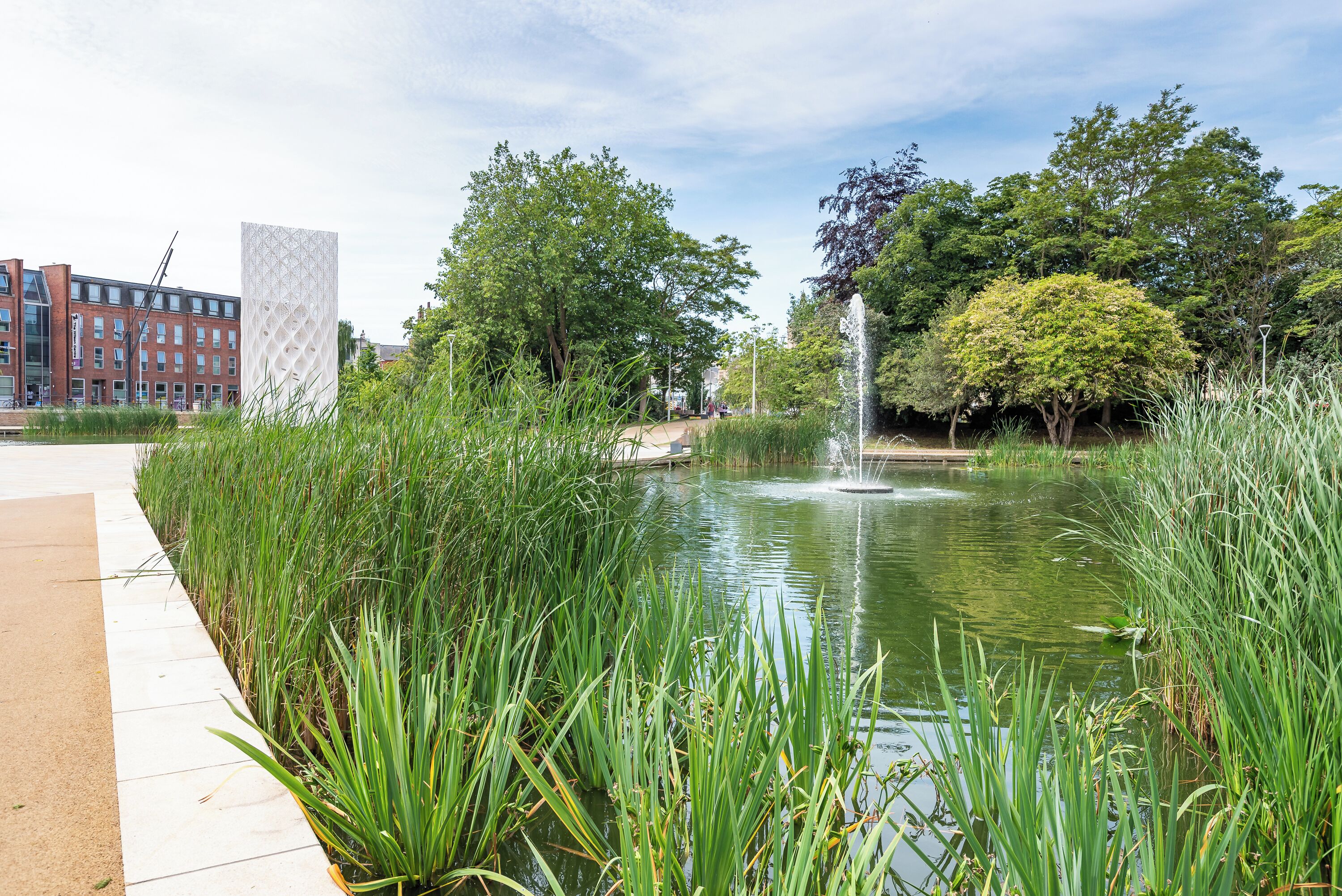 A pond with a fountain, surrounded by tall grass and trees, with a building in the background.
