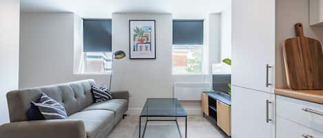 A modern living room with a sofa, a glass coffee table, and a kitchen area with cabinets and a countertop.