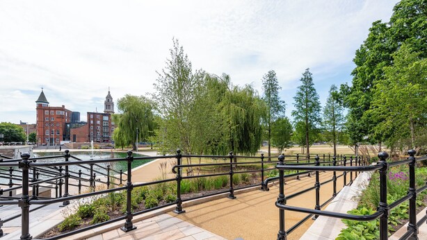 Un parco con un vialetto lastricato, un elemento d'acqua e un edificio in mattoni rossi con una torre.