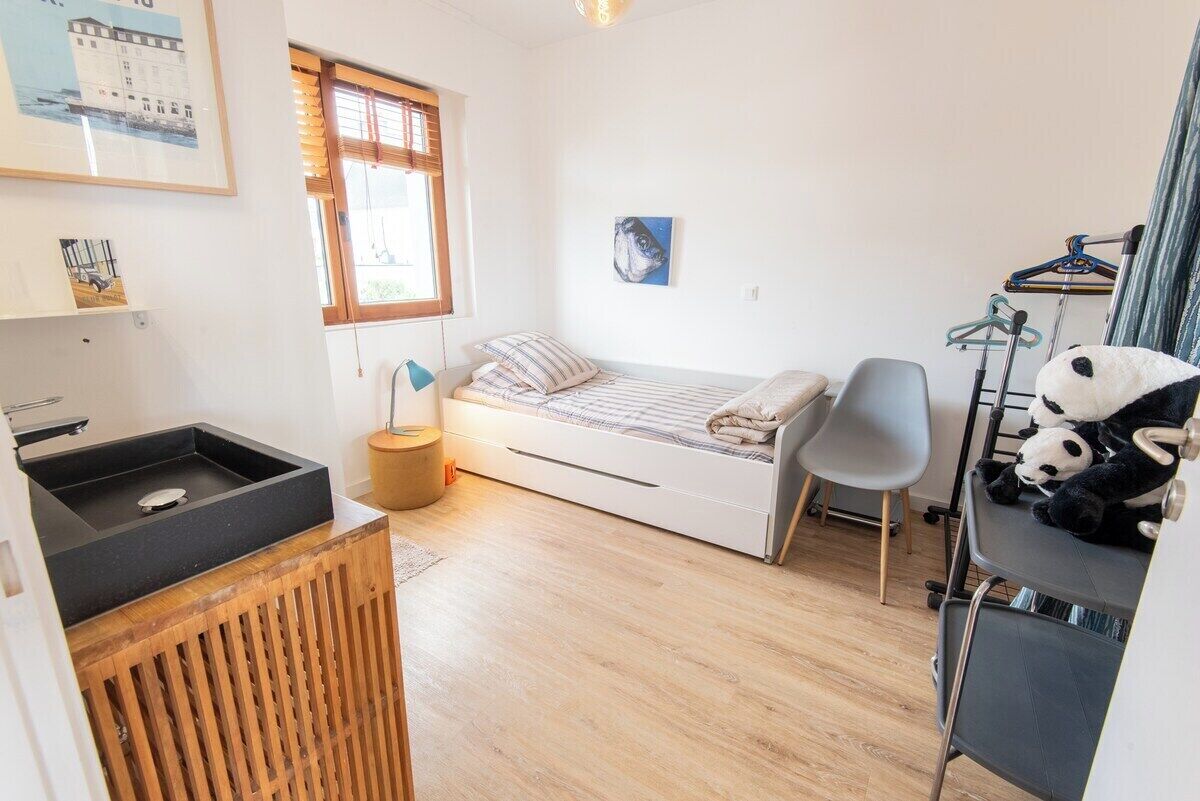 A compact room with a bed, a small desk, a chair, a window, and a wall-mounted shelf with a panda plush toy.