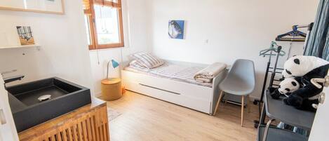 A compact room with a bed, a small desk, a chair, a window, and a wall-mounted shelf with a panda plush toy.