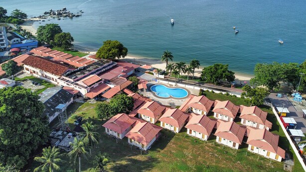 A coastal hotel with multiple bungalow-style rooms, a swimming pool, and a view of the sea and a distant island.