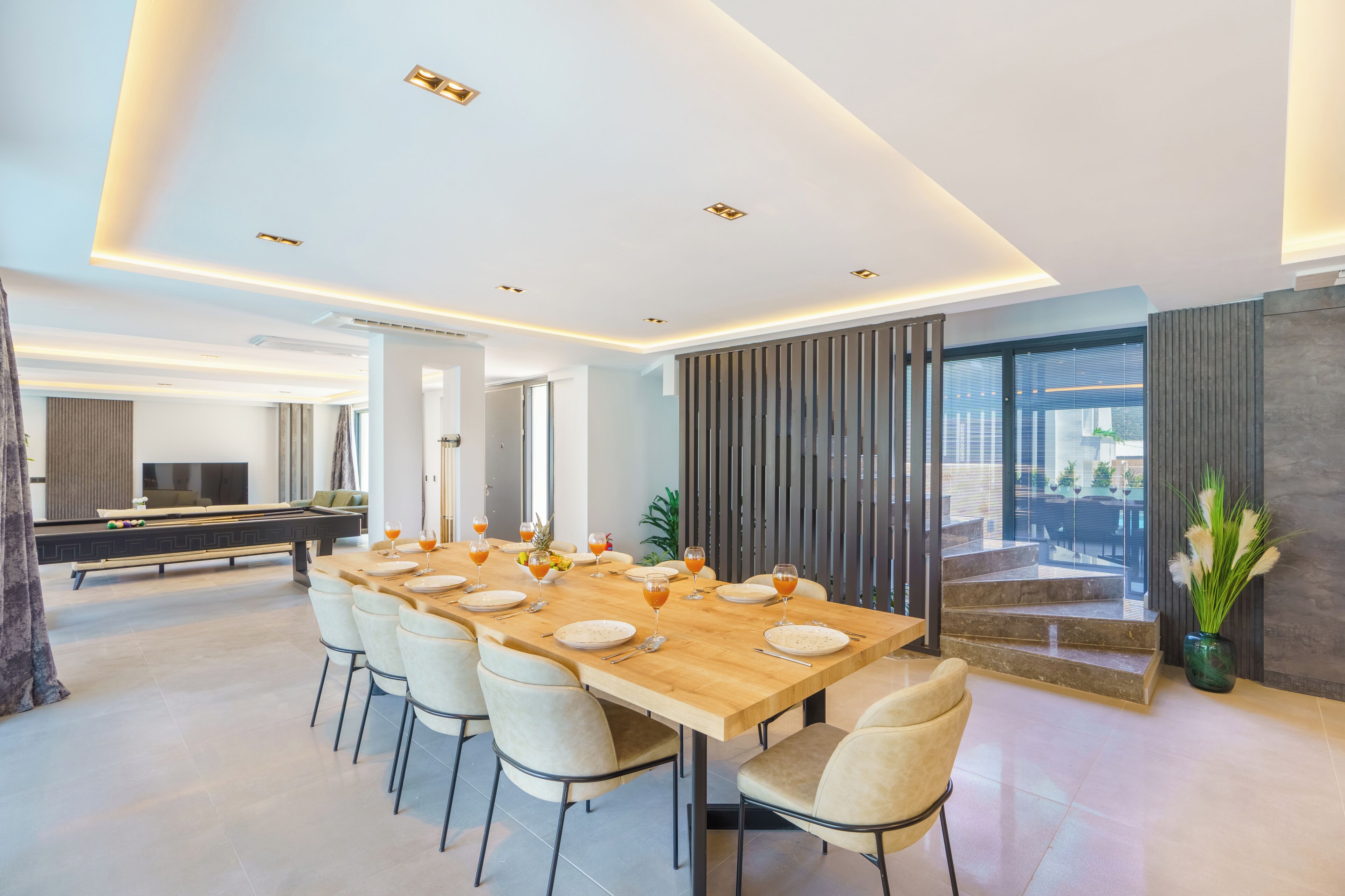 A modern dining area with a long table set for six, a pool table, and a staircase leading to an outdoor area.