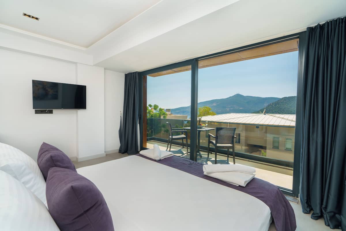 Habitación de hotel con cama, televisor de pantalla plana, balcón con mesa y sillas, y vistas a las montañas.