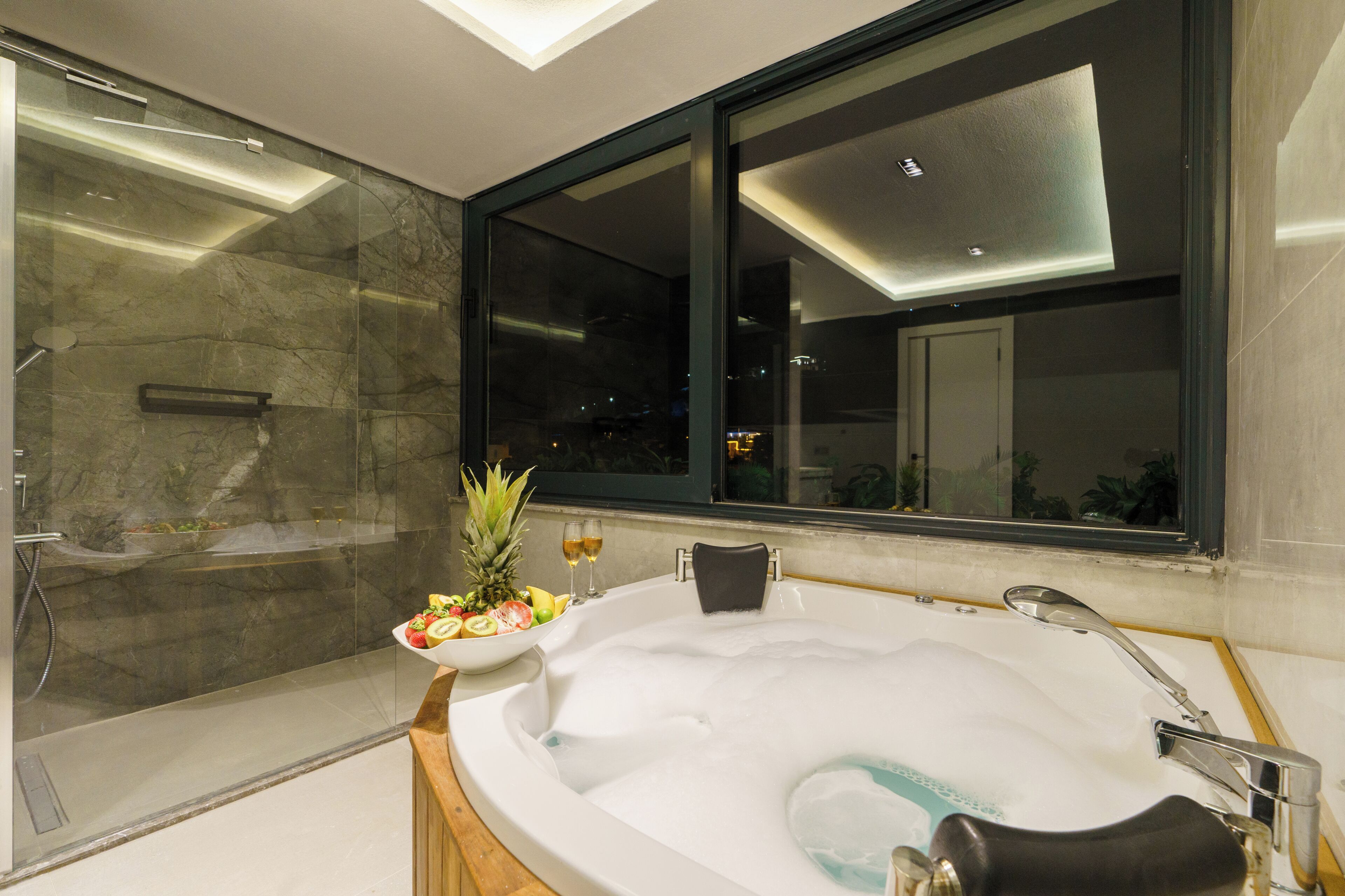 Une salle de bain moderne avec une douche à parois de verre, une grande baignoire avec des bords en bois, et un panier de fruits ainsi que deux verres de vin posés à proximité.