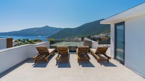 Eine Dachterrasse mit vier Holzliegen, einer Glastür und Blick auf das Meer und die Berge.