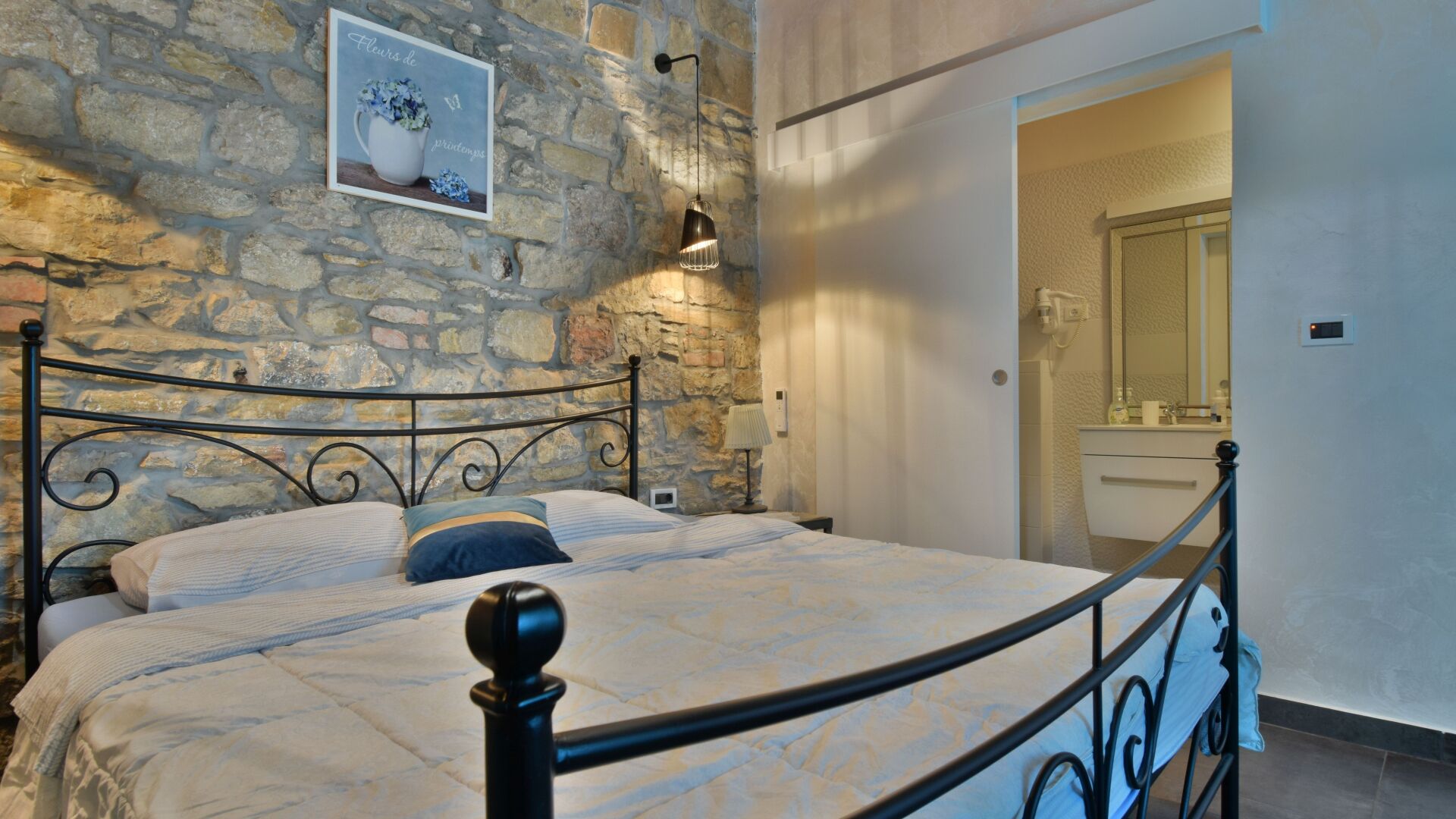 A bedroom with a stone wall, a bed with a metal frame, a bedside table, a lamp, and a mirror.