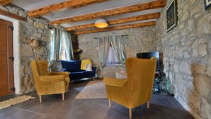 Une pièce aux murs de pierre et au plafond en bois, avec un canapé bleu, deux fauteuils jaunes et un téléviseur.