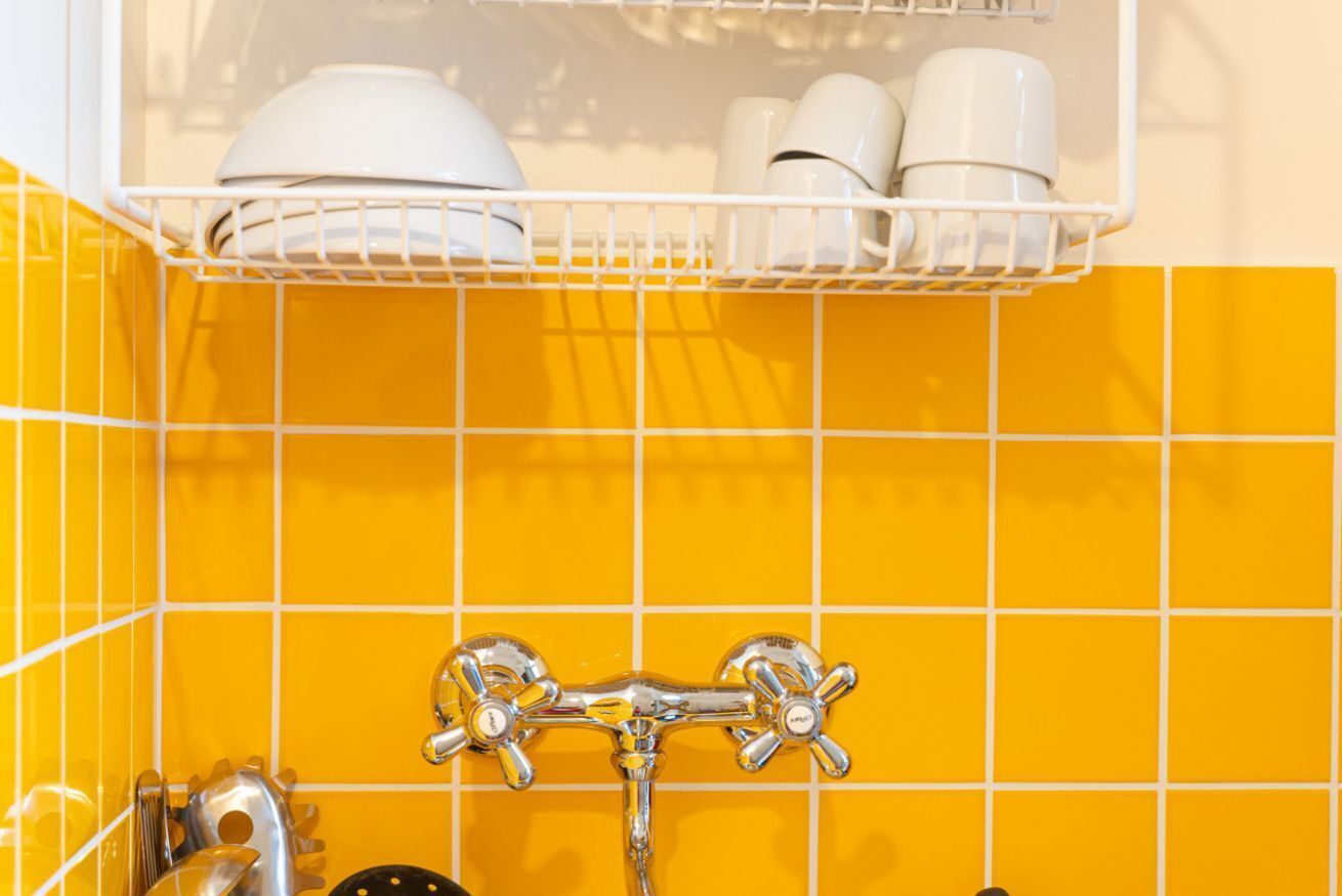 A bathroom with yellow tiles, a white shelf with cups and a bowl, and a chrome faucet.