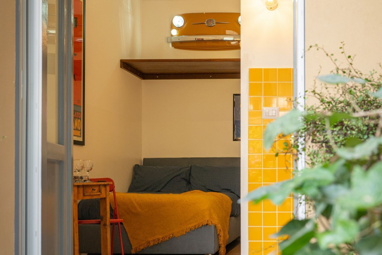 A compact room with a bed, a small table with glasses, a red chair, and a yellow-tiled wall.
