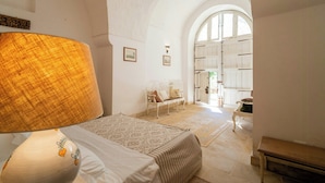 Een kamer met een bed, een lamp, een bankje en uitzicht op een keuken door een boogvormige deuropening.