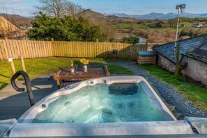 Een hottub in een achtertuin met een houten terras, een tafel met een bloevas, en uitzicht op het landschap.