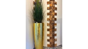 Una jardinera alta y cilíndrica de color dorado, con follaje verde y flores púrpuras, al lado de un cuadro de madera.