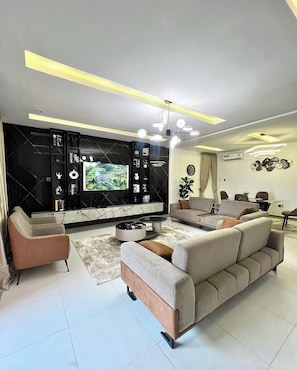 A modern living room with a large flat-screen TV, a sofa, a coffee table, and a dining area with a table and chairs. - Shiloh Manor Apartment  (Kubwa)