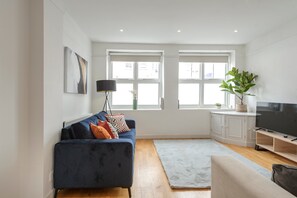Een moderne woonkamer met een blauwe bank, een grijs vloerkleed, een televisie en een groot raam.