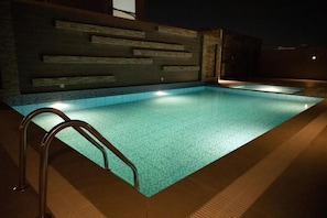 En rektangulär swimmingpool med en metallstege, omgiven av en kakelbeklädd poolterrass och en stenmur med horisontella träpaneler.