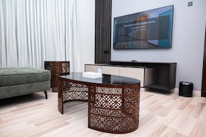 A modern hotel room with a sofa, a glass-top dining table, a TV mounted on the wall, and a decorative wooden partition. - Jadeel Hotel (Al Khobar)