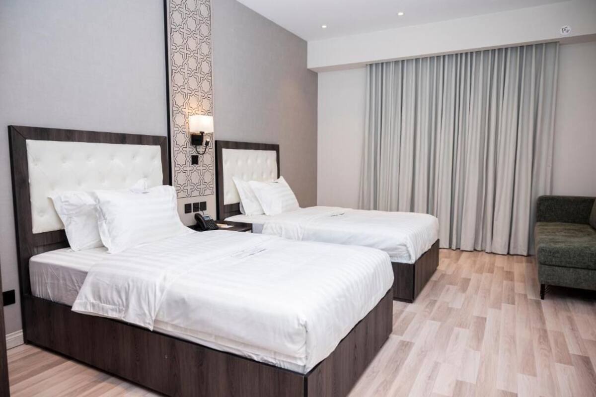 Ett hotellrum med två sängar, en grön fåtölj, en mönstrad vägg och ett stort fönster med gardiner.