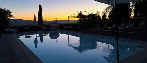 A swimming pool with a clear view of the sunset in the background, surrounded by lounge chairs and a pergola.