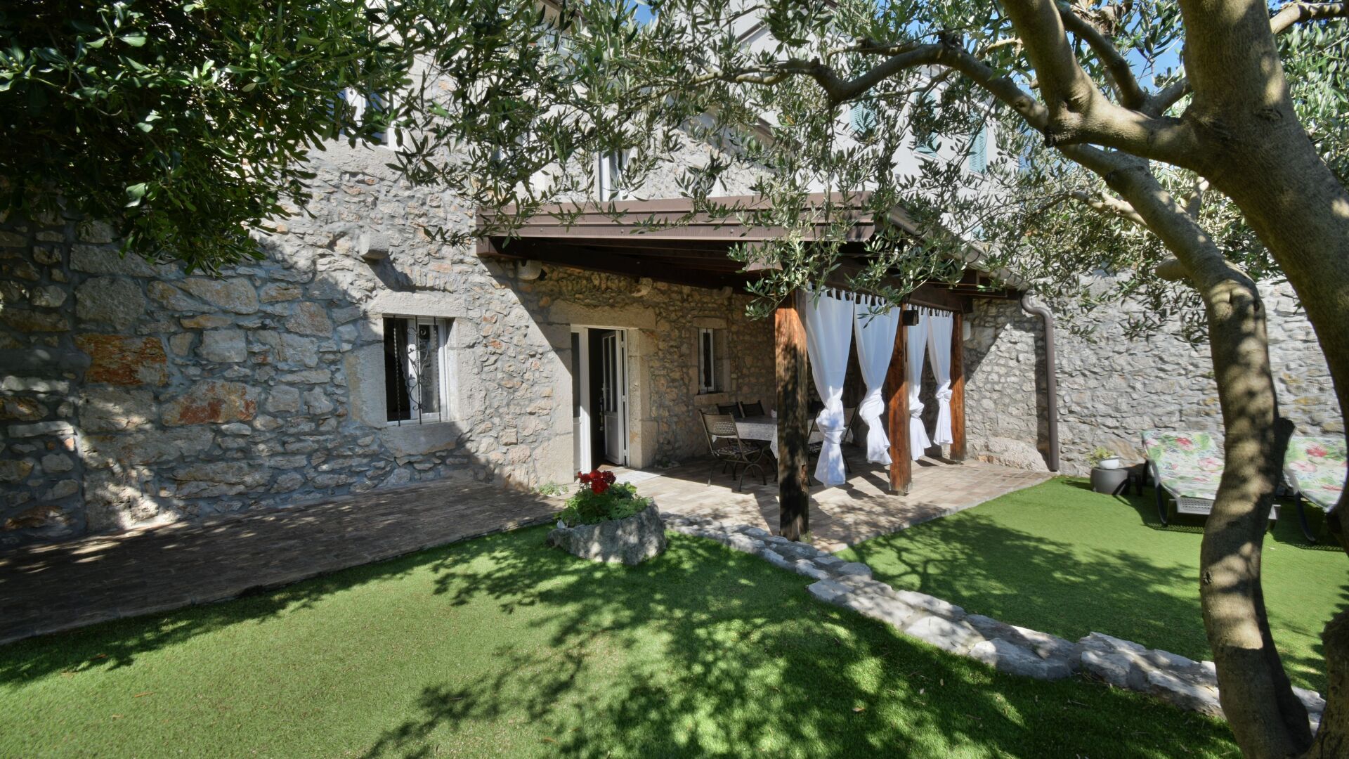 Un edificio in pietra con un'area salotto esterna coperta, una piccola finestra e un giardino con erba sintetica.
