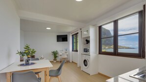 Eine moderne Küche mit Waschmaschine, einem Essbereich mit Stühlen, einem an der Wand befestigten Fernseher und einem großen Fenster mit Blick auf eine Küstenlandschaft.