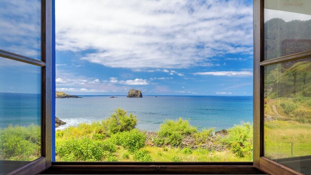 Blick auf die Küste durch ein Fenster, mit blauem Himmel, Meer und fernen Felsen.