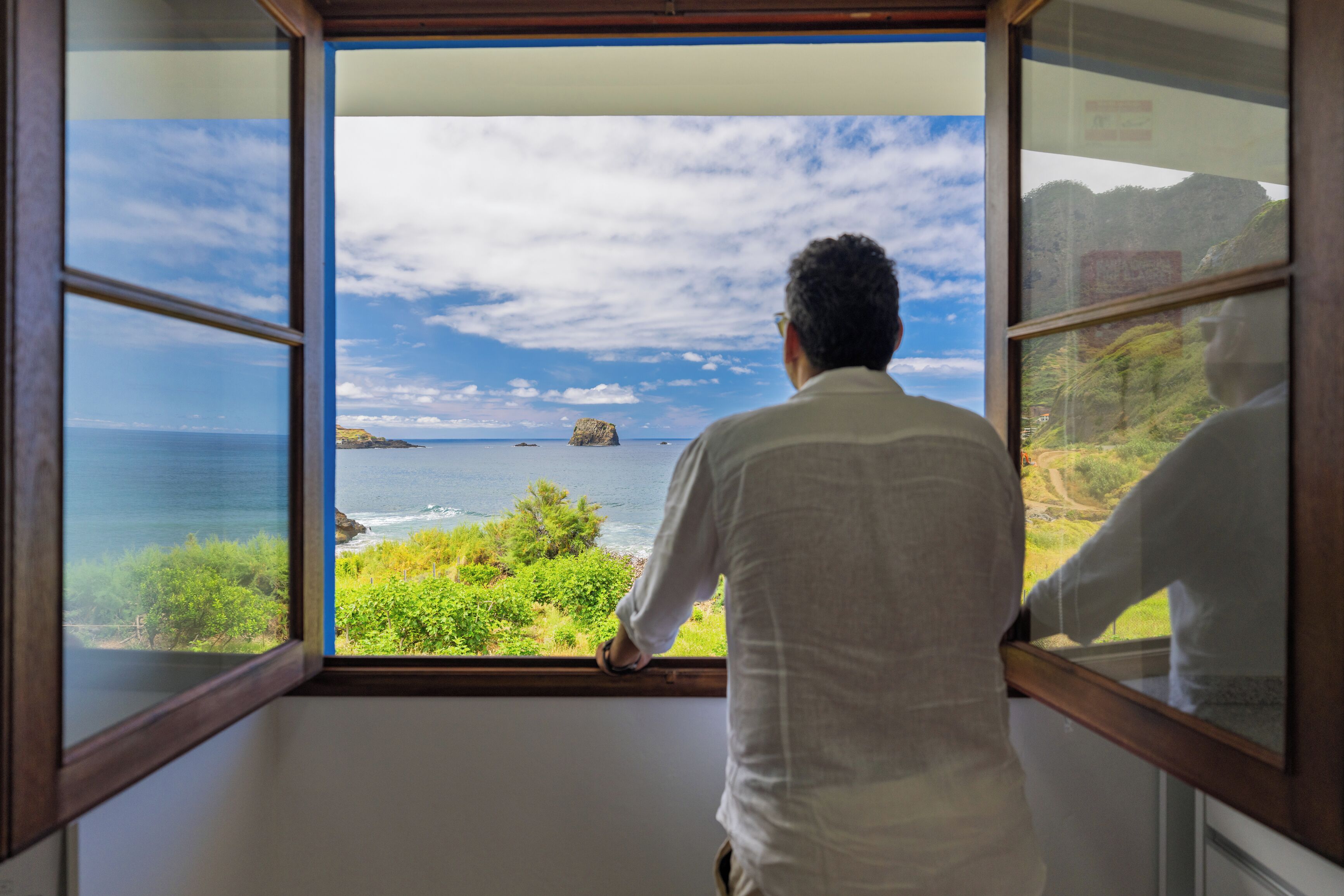 A man looking out of a window with a view of the ocean and greenery.