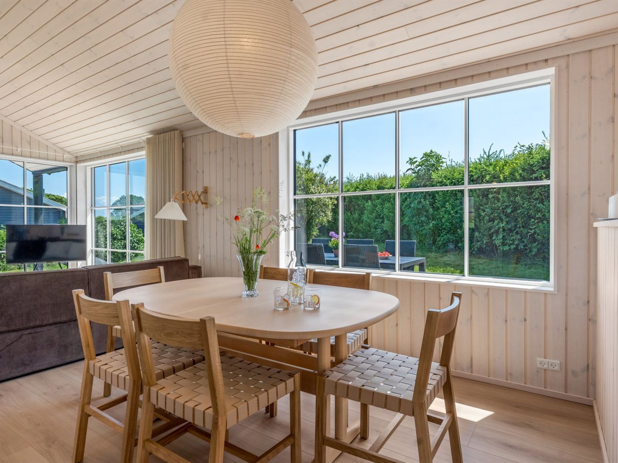 A dining area with a round table, wooden chairs, a vase with flowers, and a large window offering a view of greenery.