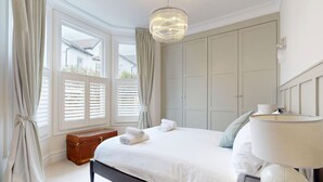 A bedroom with a bed, bedside lamp, curtains, a window with shutters, a chest of drawers, and a wardrobe. - Prime 3BR Flat With Private Patio-Walk to Tube (Richmond)