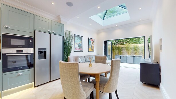 A modern kitchen with a dining area, featuring a built-in oven, refrigerator, and a skylight above the dining table. - Prime 3BR Flat With Private Patio-Walk to Tube (Richmond)