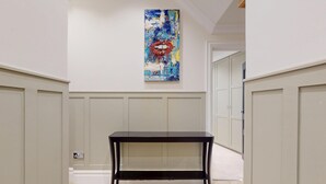 A hallway with a black console table, a white wall with a blue abstract painting, and a door with a white panel. - Prime 3BR Flat With Private Patio-Walk to Tube (Richmond)